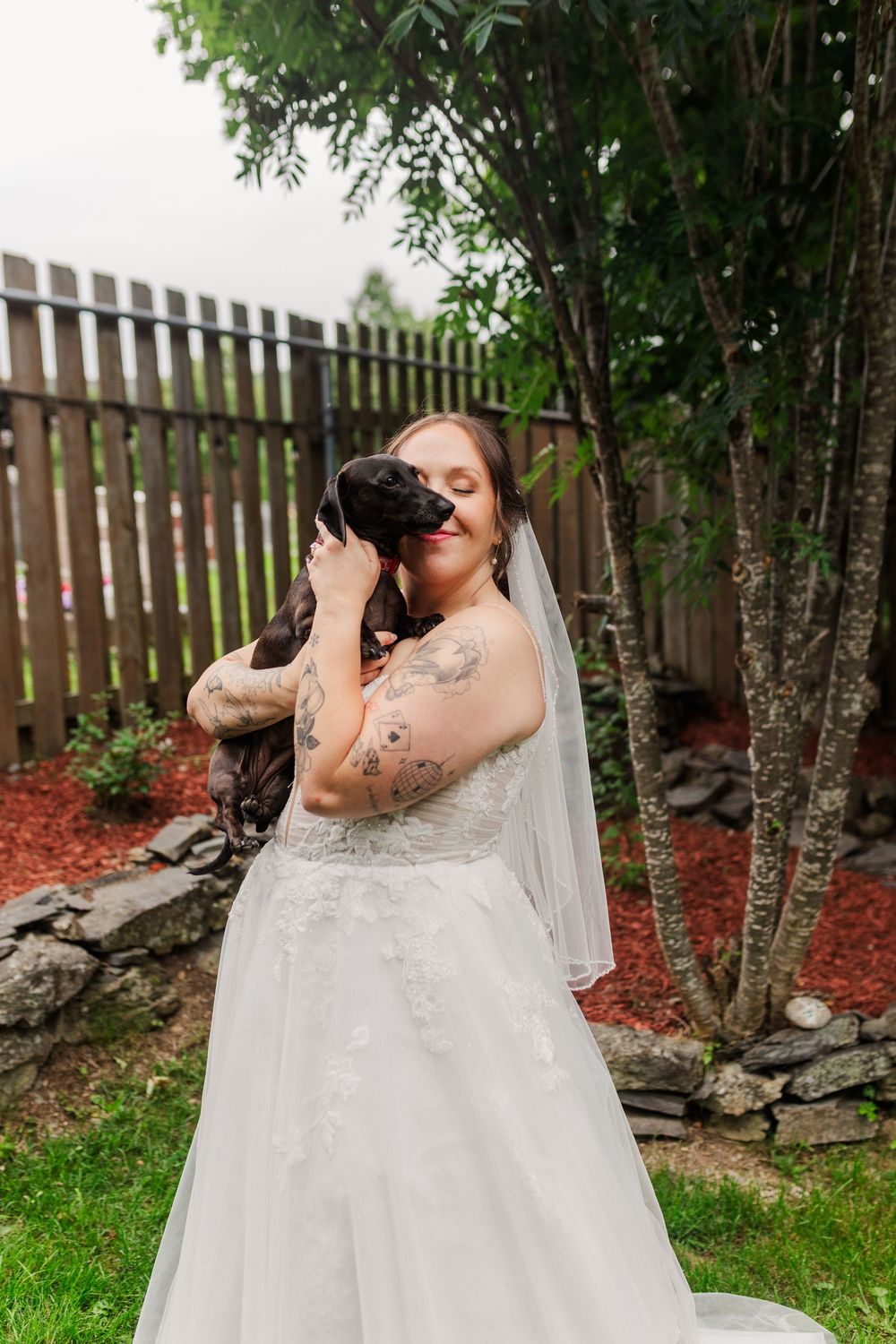 Tattooed bride in white gown and veil cuddles small black dog in garden with wooden fence backdrop.
