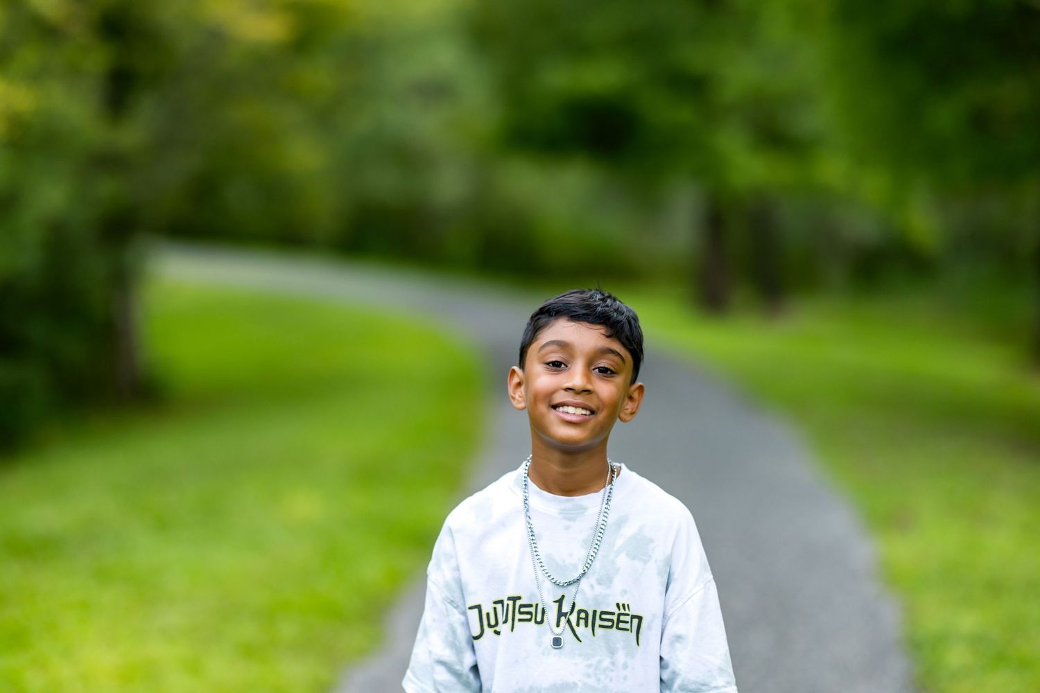 Smiling young boy wearing a Jujutsu Kaisen sweatshirt standing on a park path surrounded by green trees.
