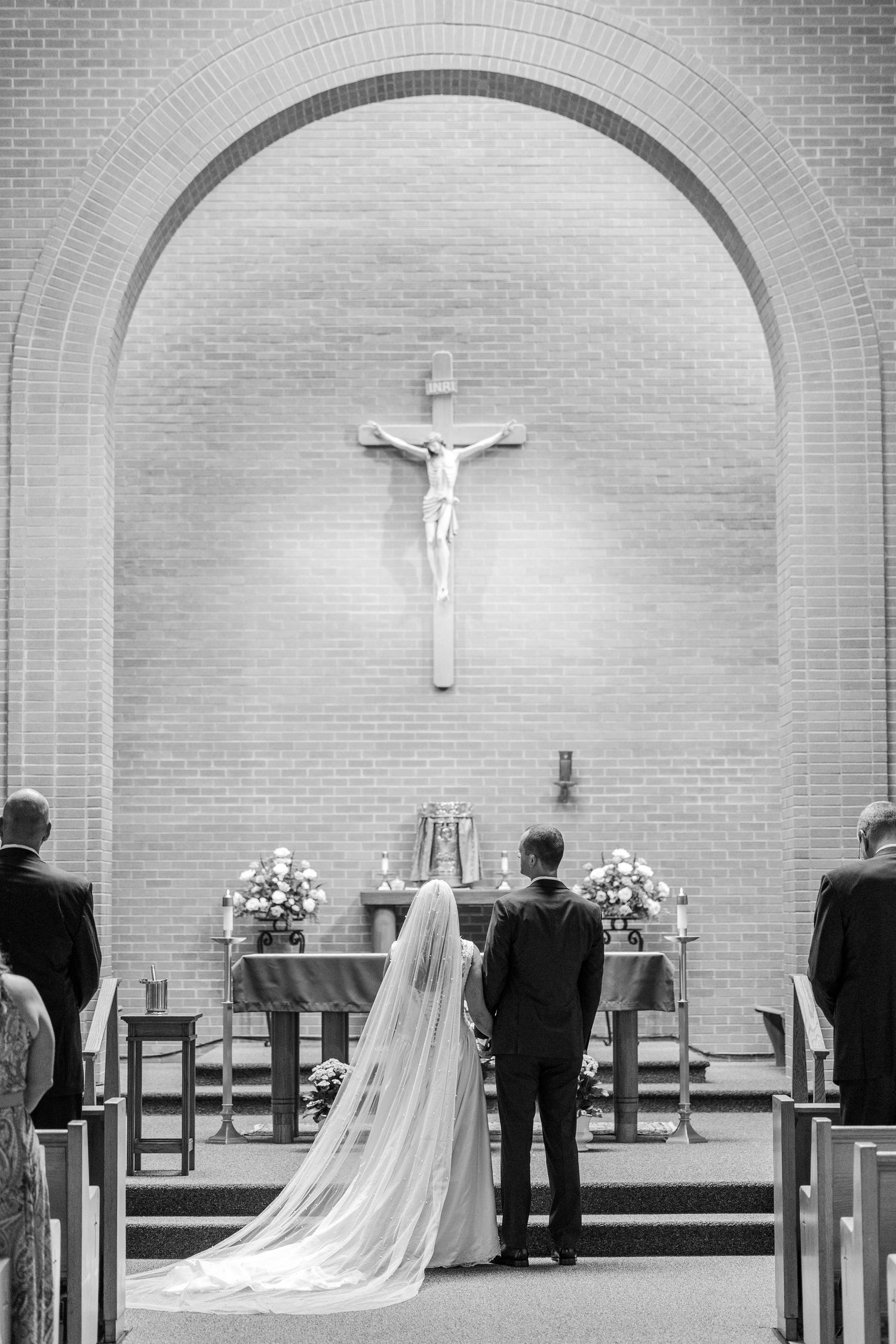 Black and white photo of bride and groom standing at church altar beneath crucifix during Catholic wedding ceremony.
