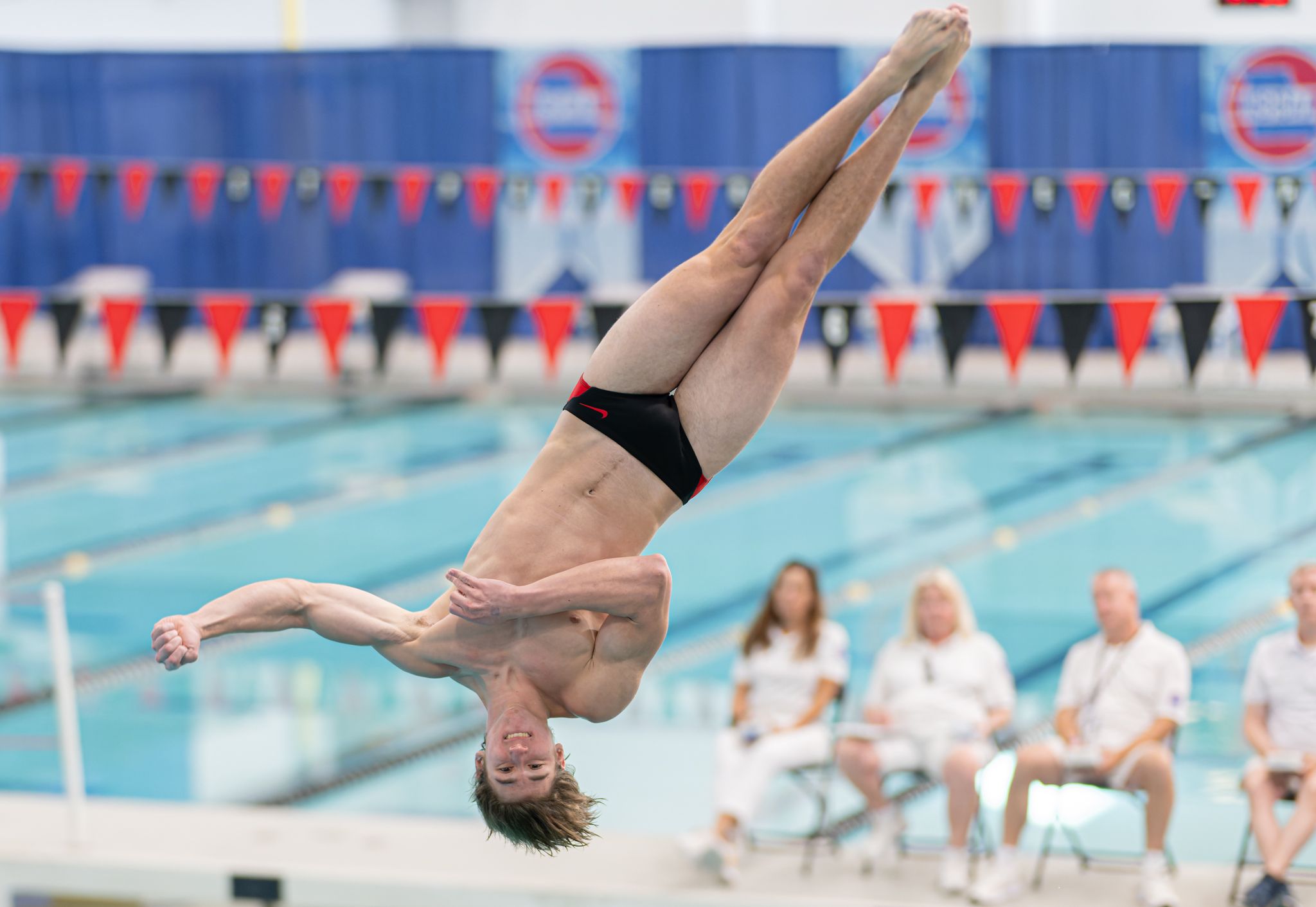 Male diver performing a backflip over an indoor swimming pool during a competitive diving event.