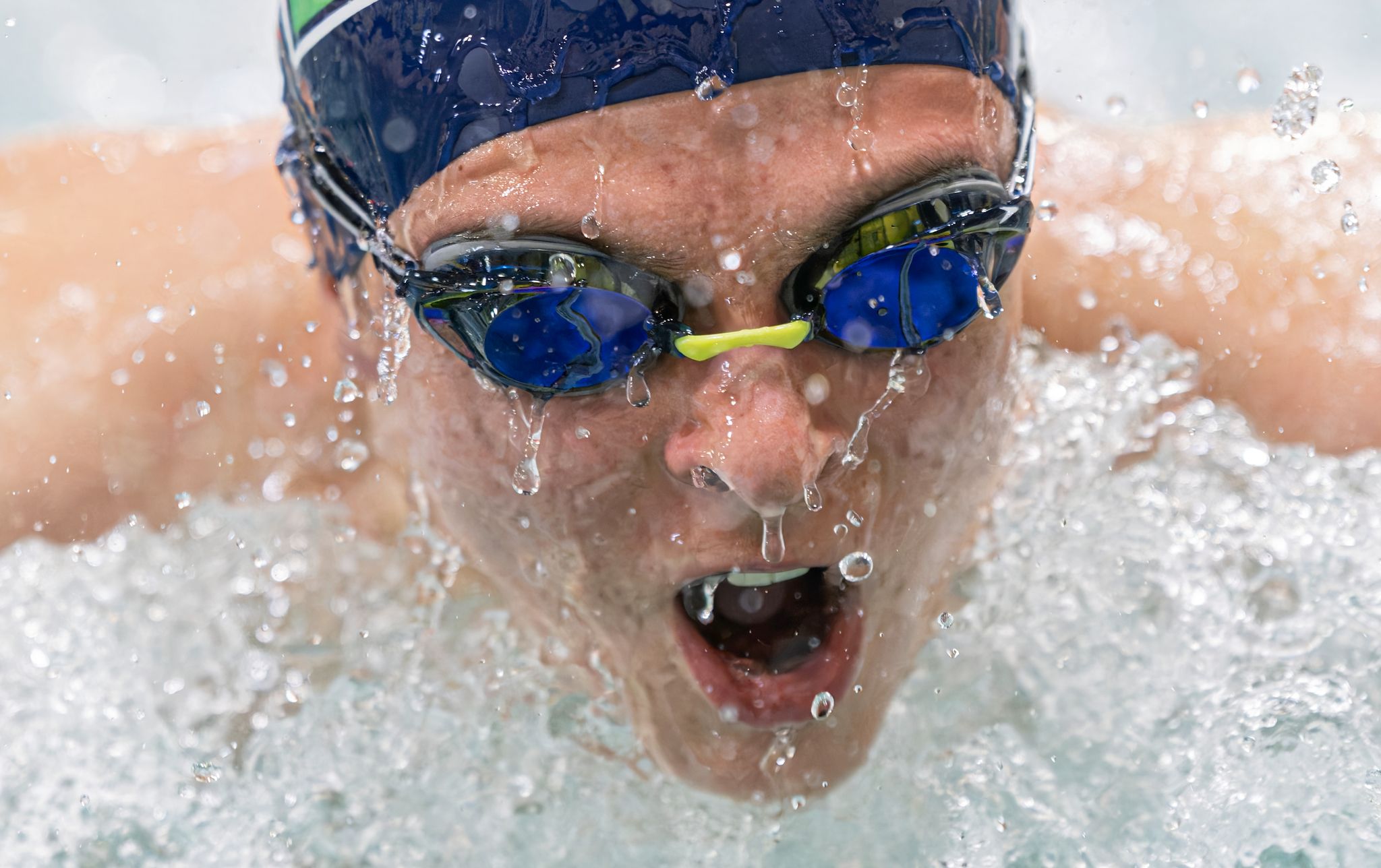 Close-up of a competitive swimmer wearing blue goggles and cap, splashing through water during a race.
