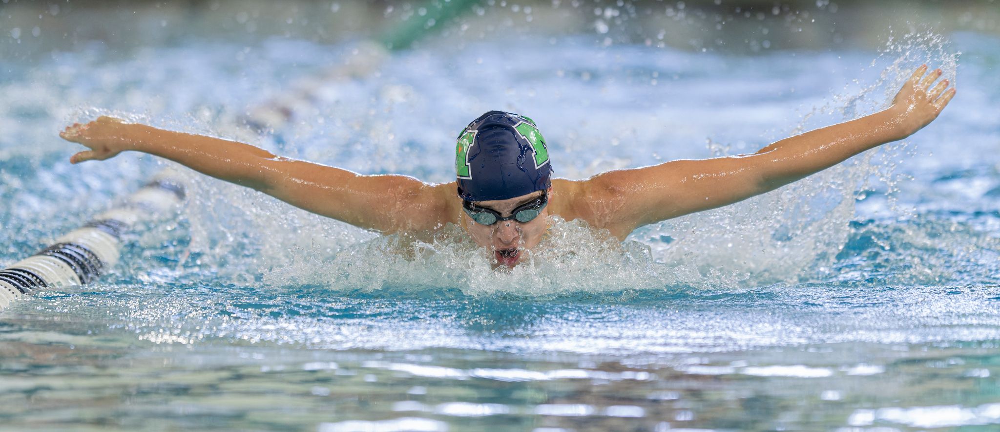 Competitive swimmer performing butterfly stroke in pool, wearing green cap and goggles during a swim meet.