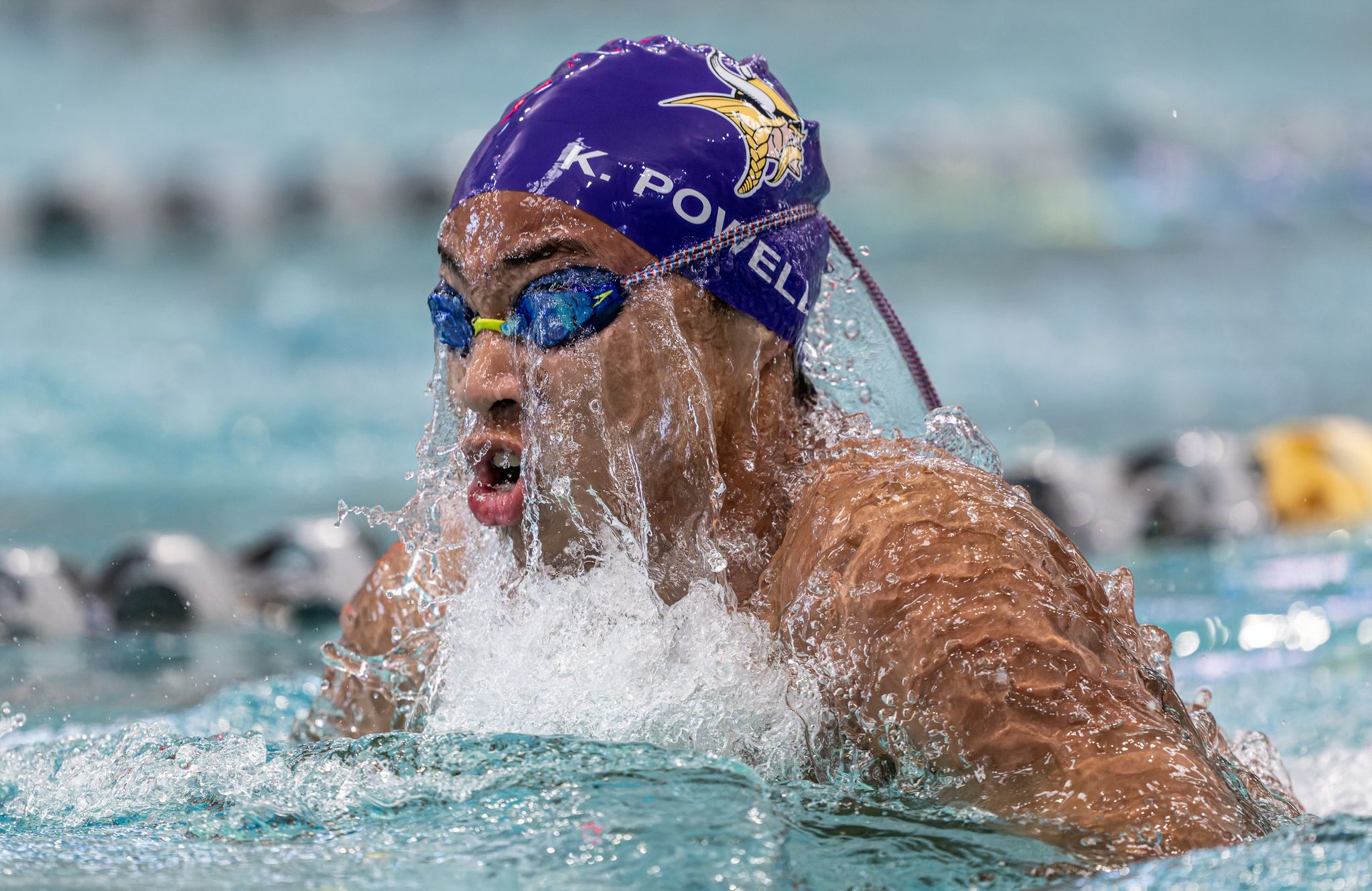 Swimmer K. Powell wearing purple cap and goggles competes in breaststroke race at indoor swimming pool.