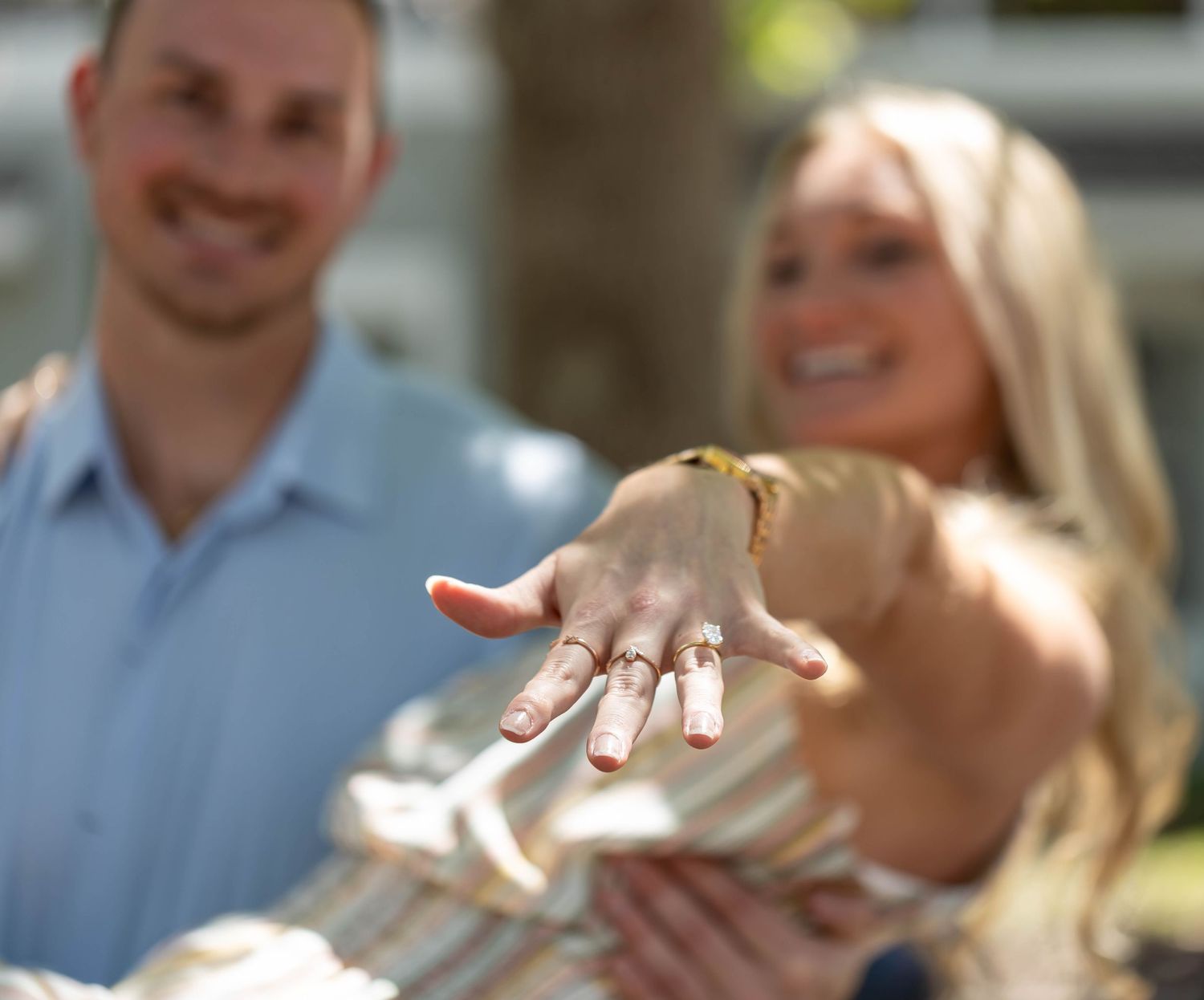 A newly engaged couple showing off the ring in bright sunshine outdoors.