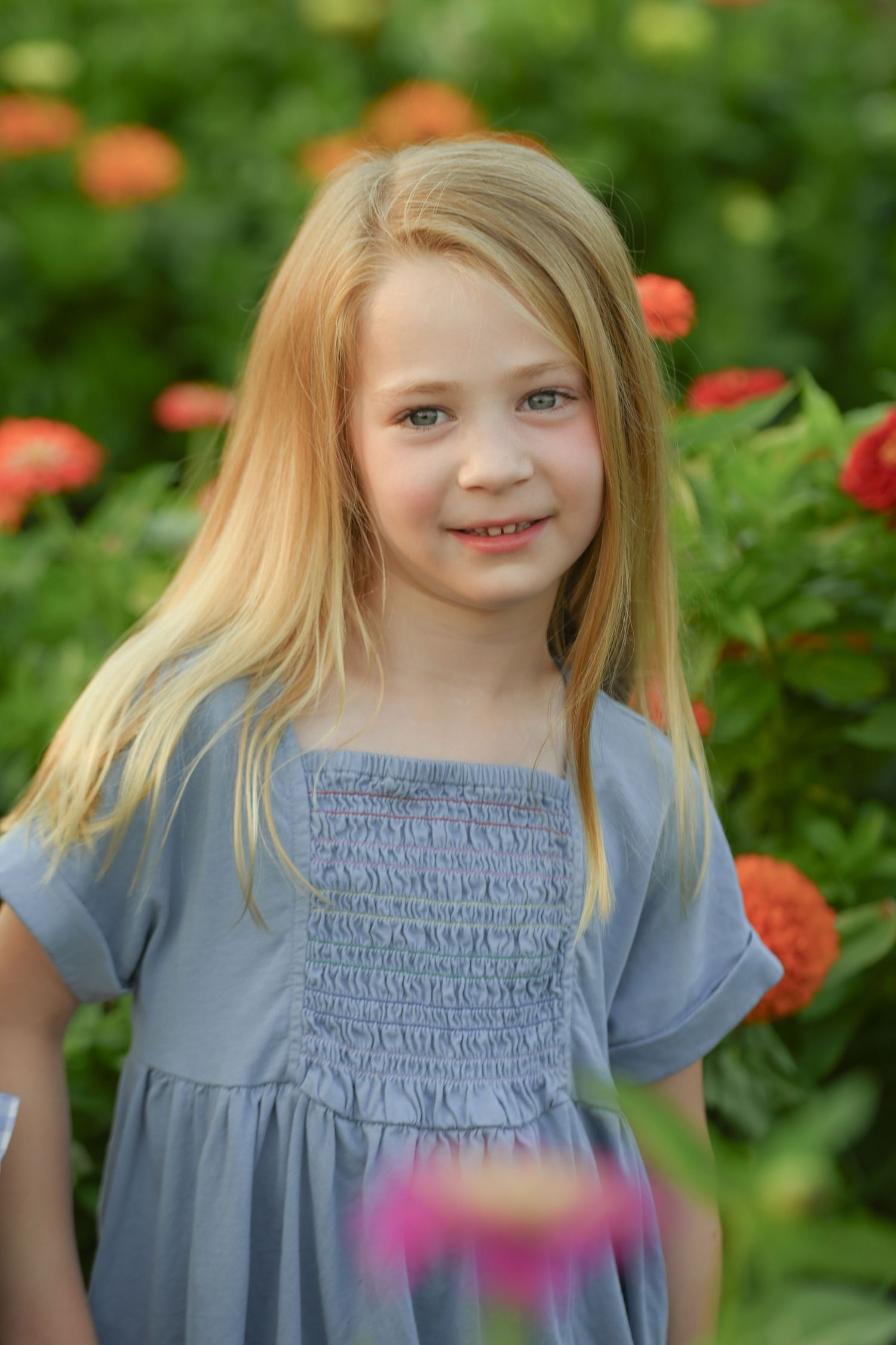 Young blonde girl in a blue smocked dress smiling in a garden with colorful zinnia flowers in the background.