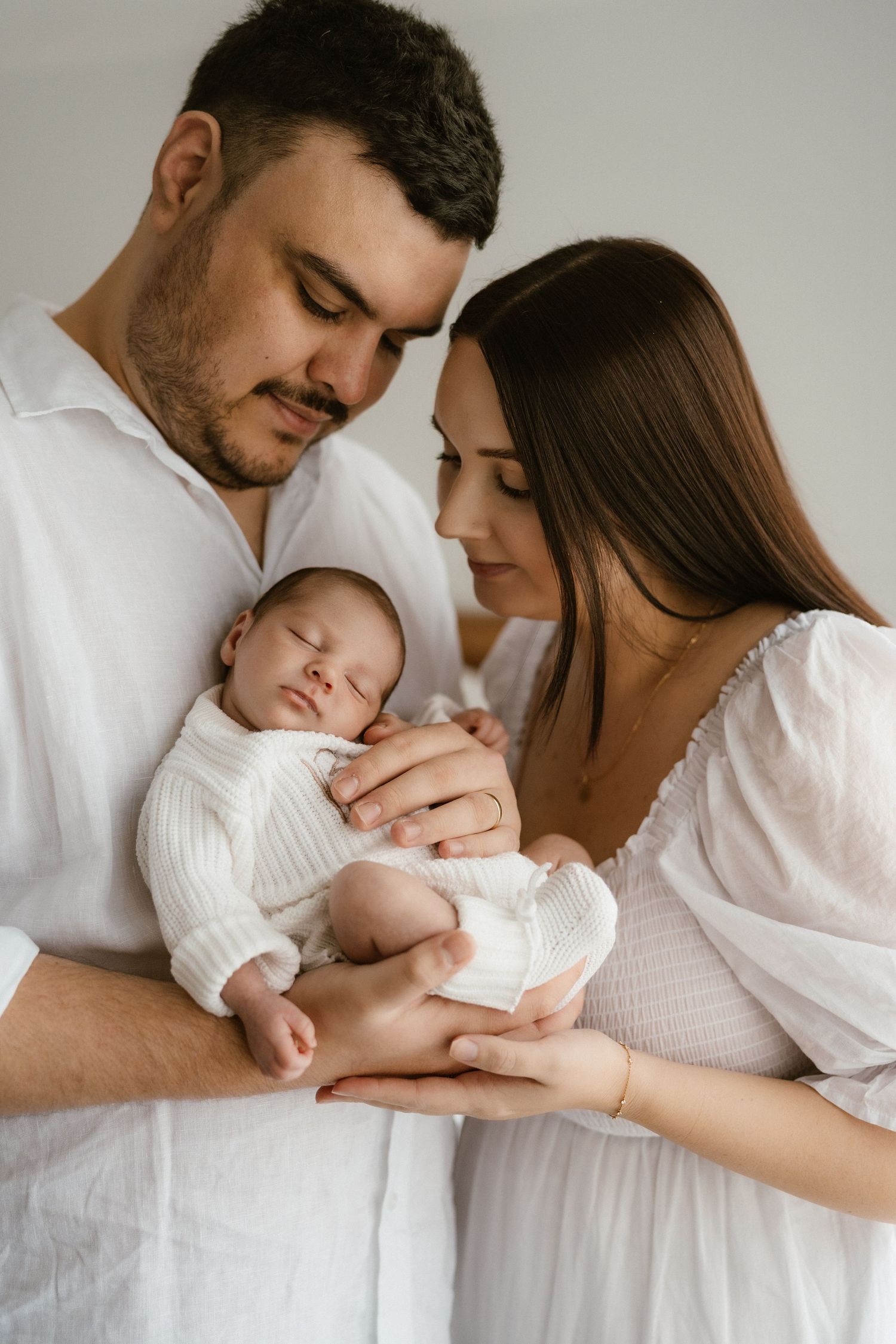 A young couple in white clothing tenderly gazes at their sleeping newborn baby cradled in their arms.