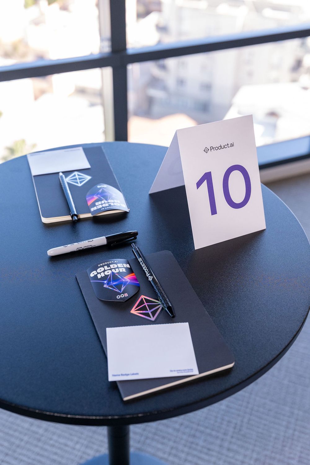 Round black table with number 10 sign, notebooks, stickers, and pens at a Product.ai event.