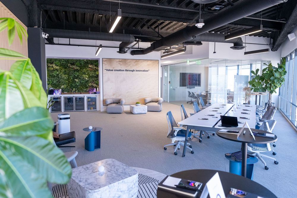 Modern open-plan office with living green wall, long conference table, ergonomic chairs, and casual lounge seating area.