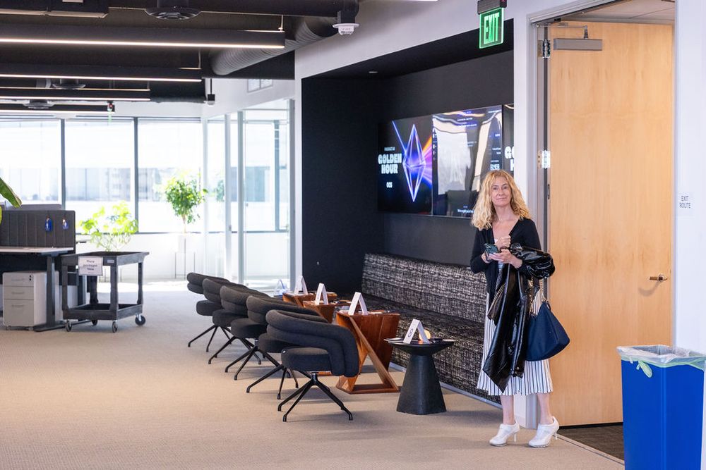 Woman standing in modern office space with dark accent wall, TV display, and open workspace area.
