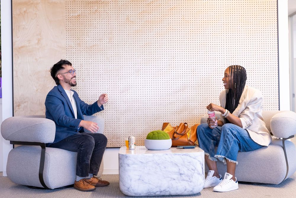 Two colleagues having a casual conversation seated in modern lounge chairs around a marble coffee table in a bright office.