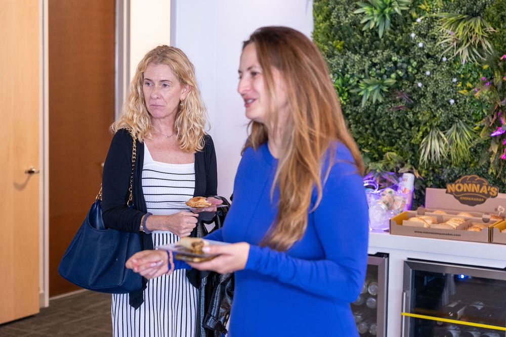 Two women socializing at a catered office event, one in a blue top and another in a striped dress holding food.