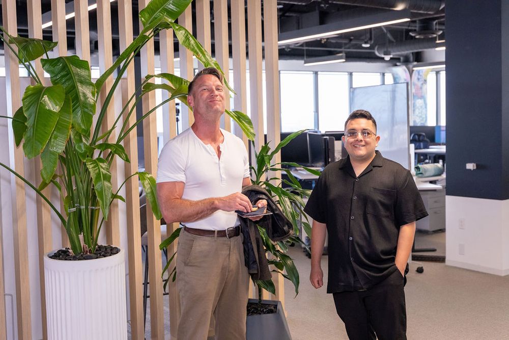 Two men standing in a modern office space with tropical plants and open workspace in the background.