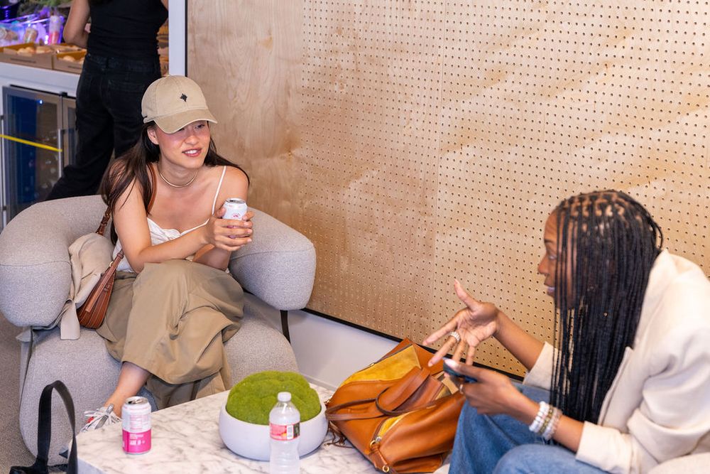 Two women socializing in a modern lounge, one holding a drink, with a handbag and beverages on the table.
