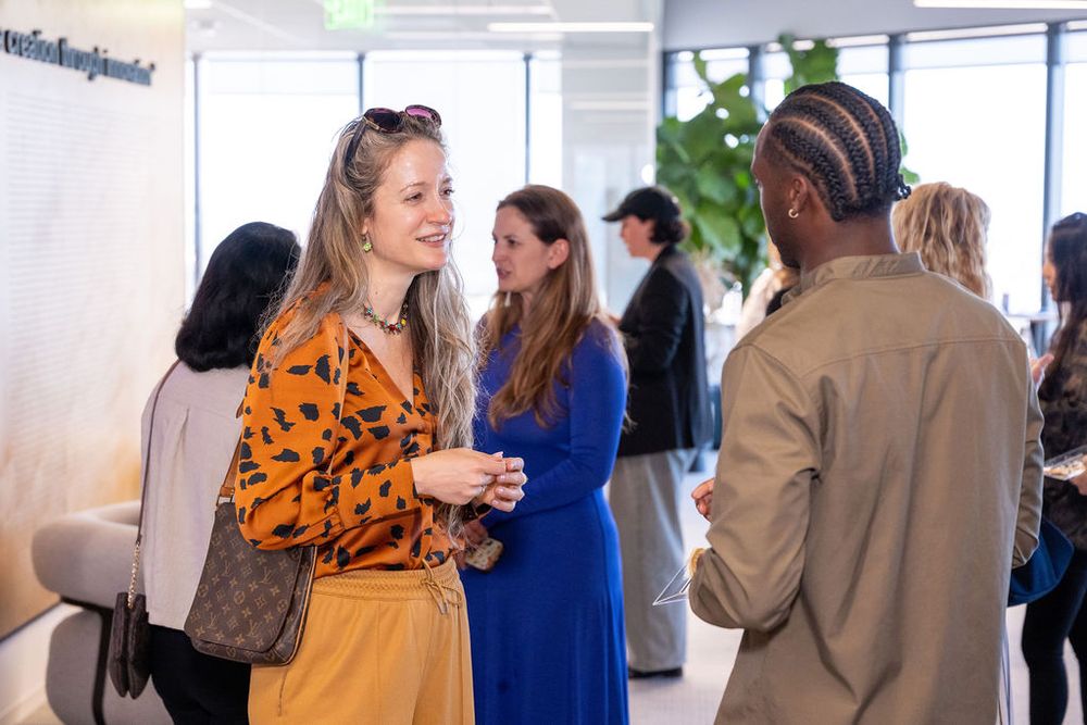 Professionals networking at a modern office event, with a woman in an orange floral top conversing with colleagues.