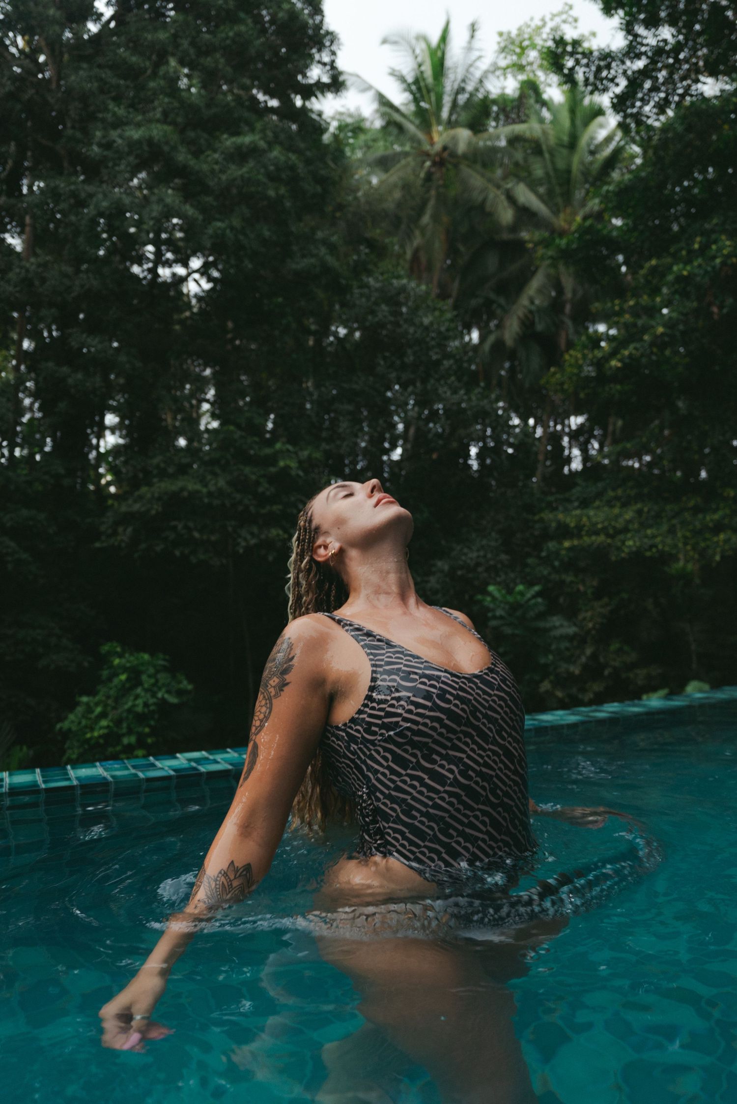 Woman in patterned swimsuit emerging from tropical pool, wet hair back, tattoo visible, lush green trees background.