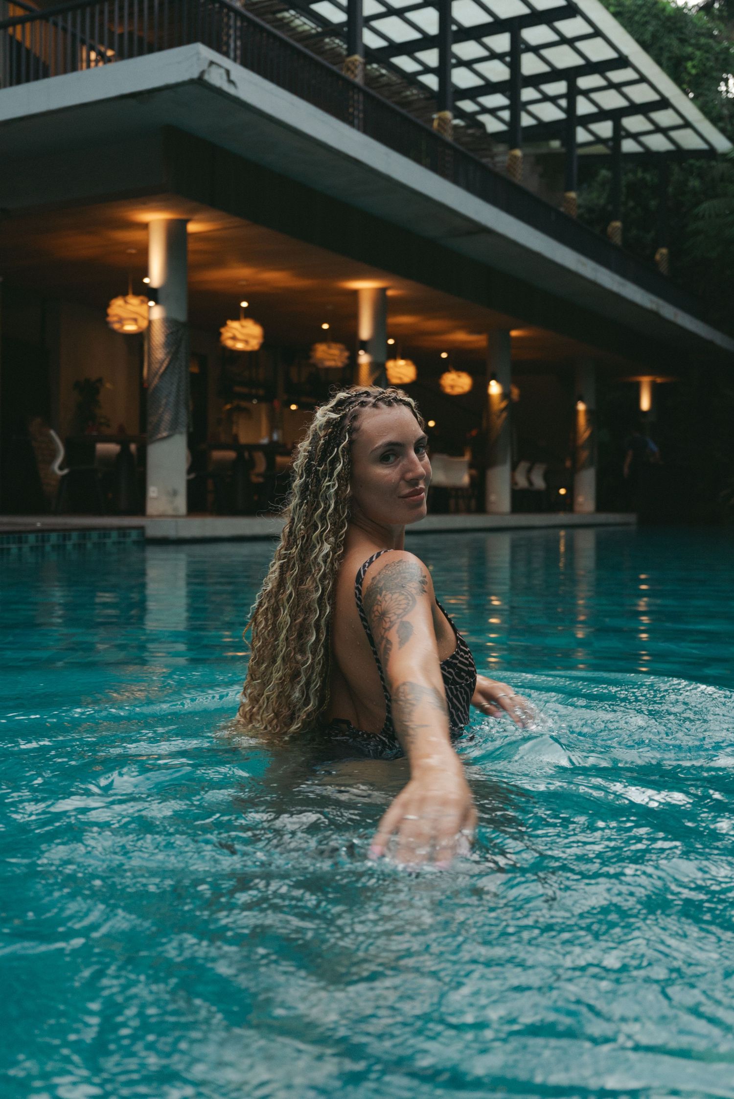 Woman with long curly hair swimming in a resort pool at dusk, with illuminated bar area in background.