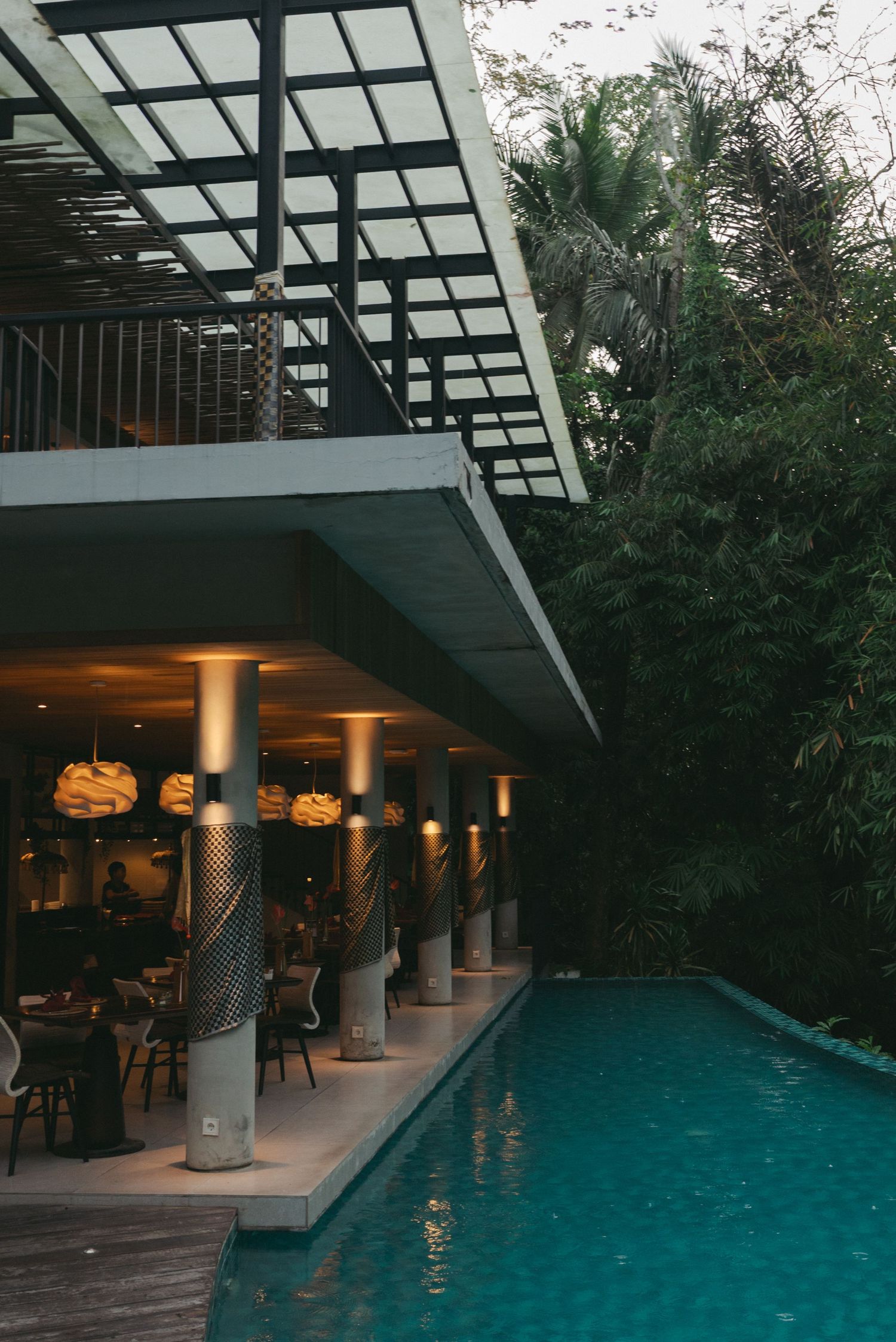 Modern resort pool area with illuminated columns, outdoor dining, and lush tropical greenery at dusk.