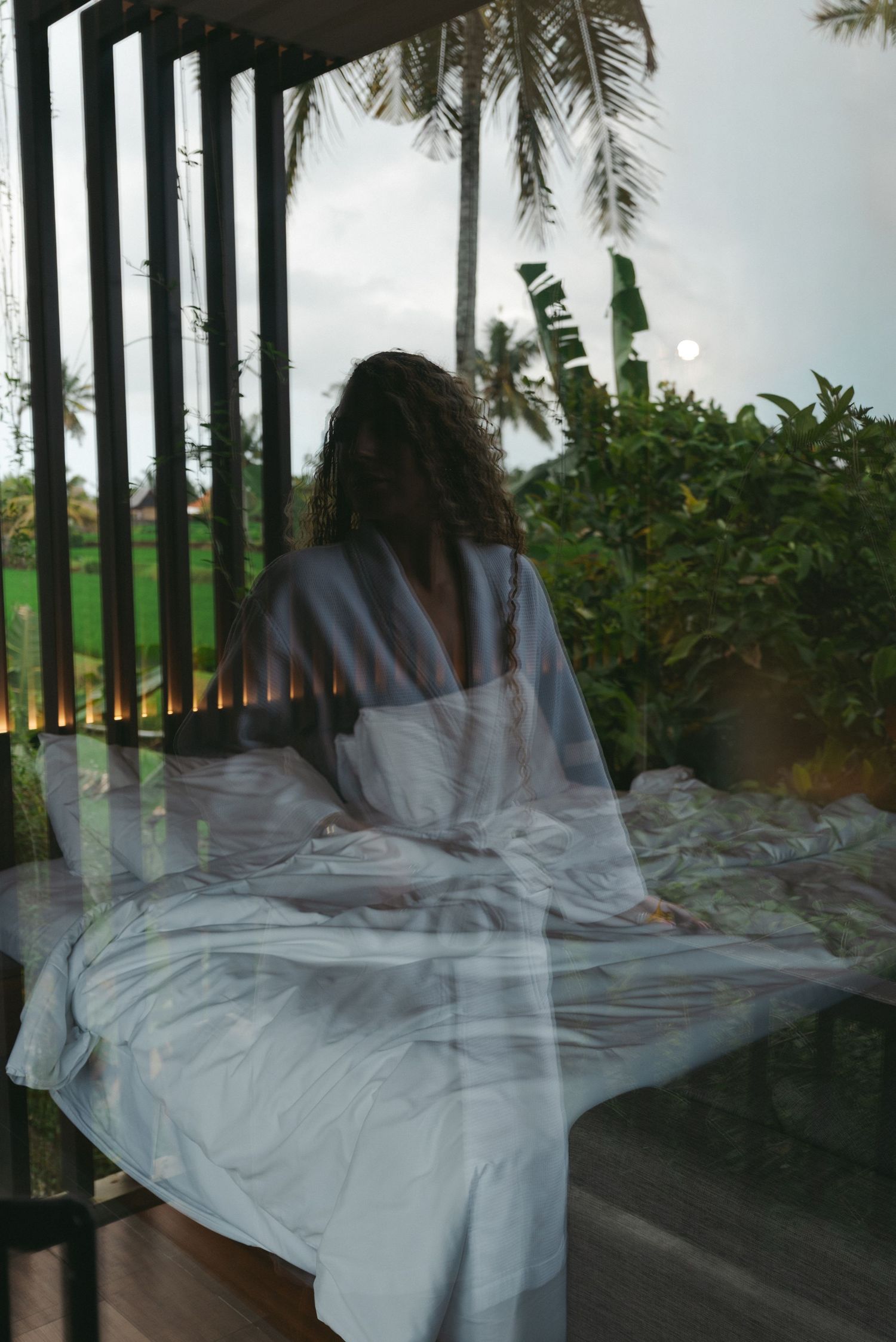 Woman in white dress sitting on a porch, viewed through glass with tropical palm trees in background.