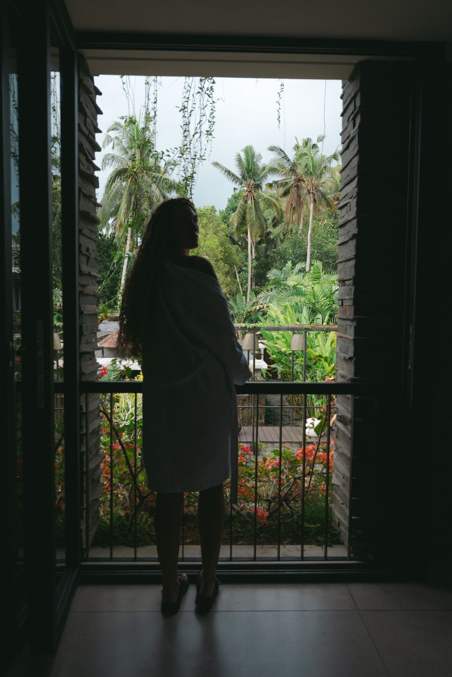 Silhouette of a woman in a white robe standing on a tropical balcony overlooking lush palm trees and colorful flowers.