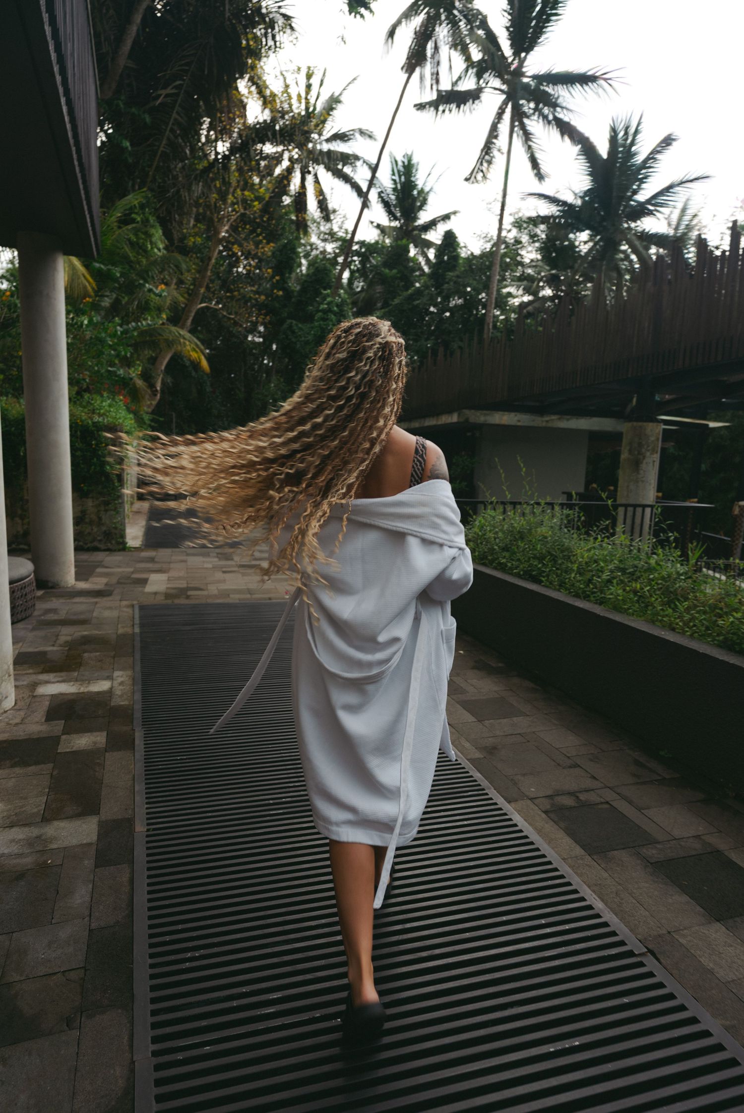 Woman in white robe with braided hair walking along tropical resort pathway surrounded by palm trees.