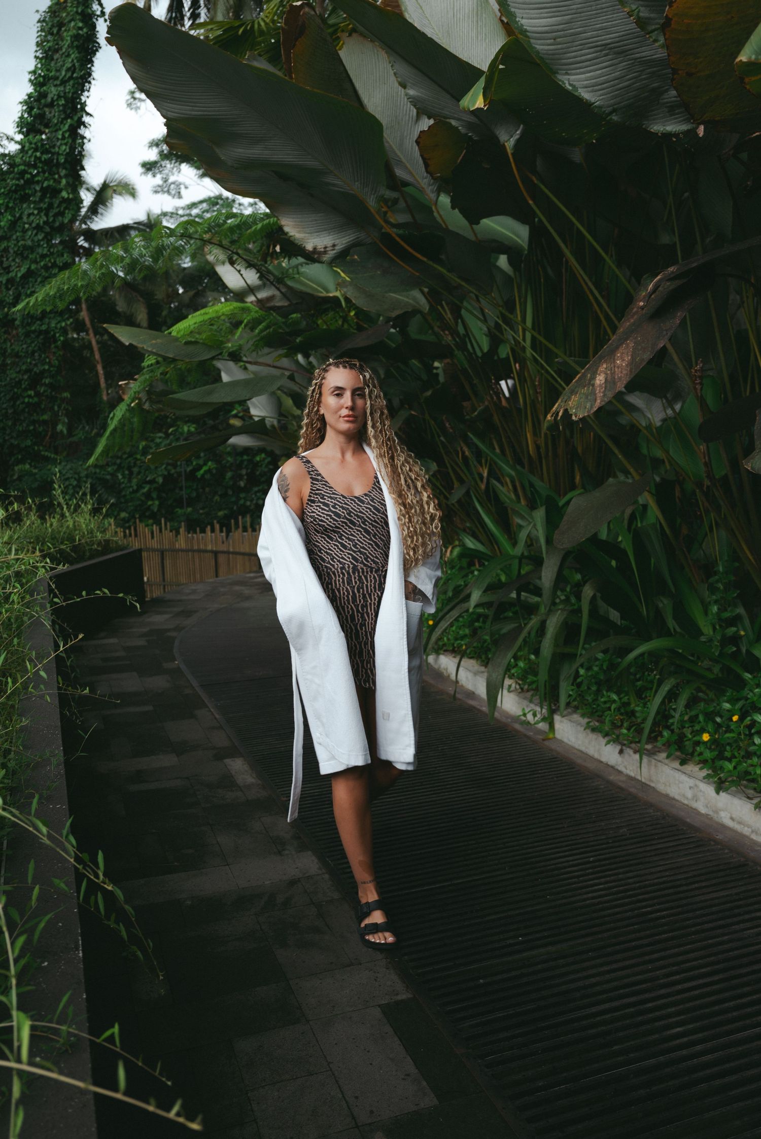 Woman with curly hair in animal print dress and white jacket walks tropical garden path.