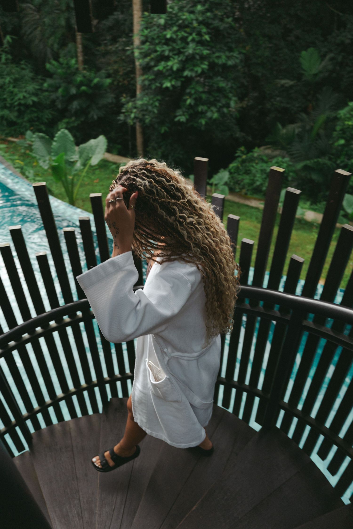 Woman in white robe with curly hair stands on wooden balcony overlooking lush tropical garden and pool.