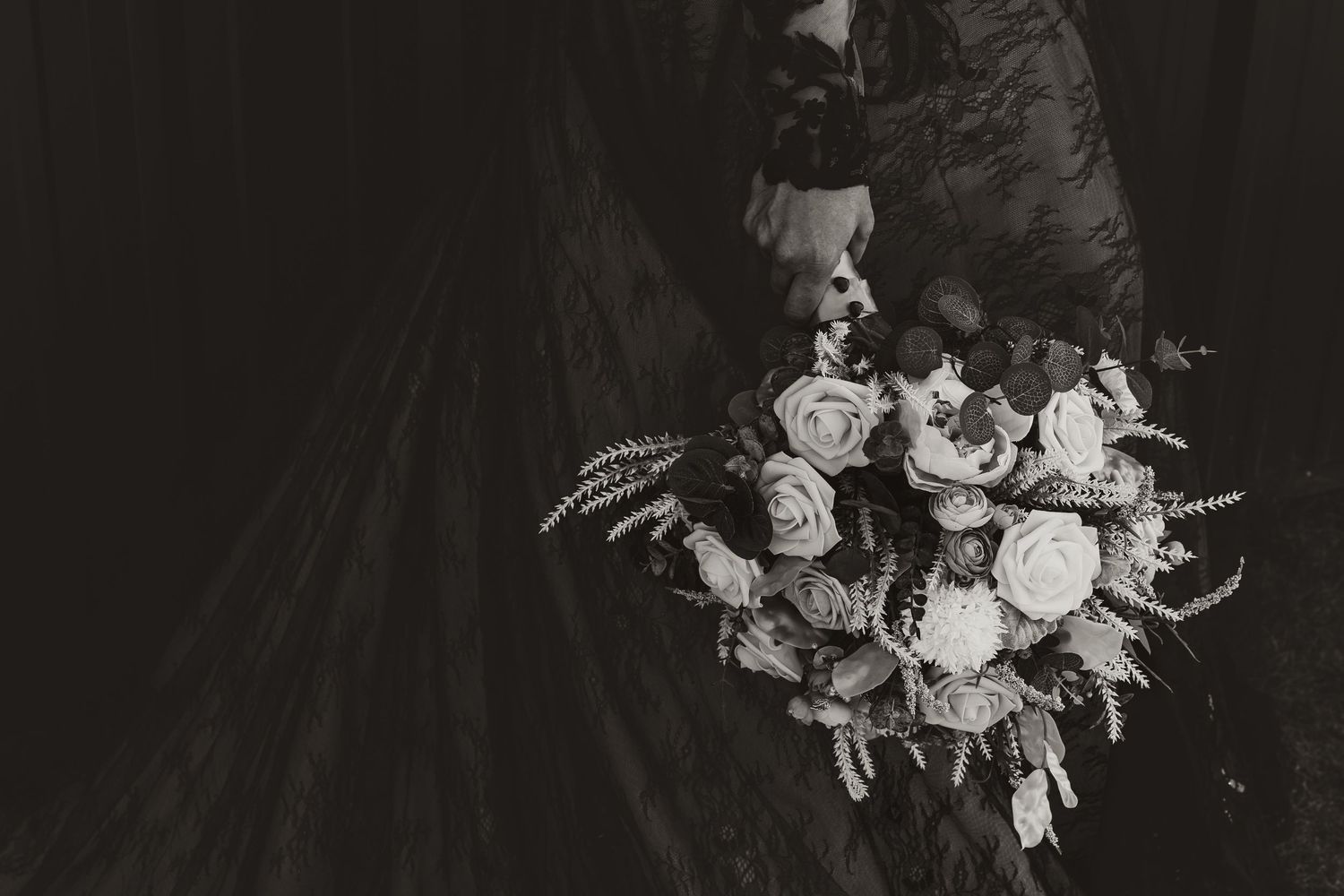 Black and white close-up of a bride holding a lush rose and greenery bouquet against a dark gown.