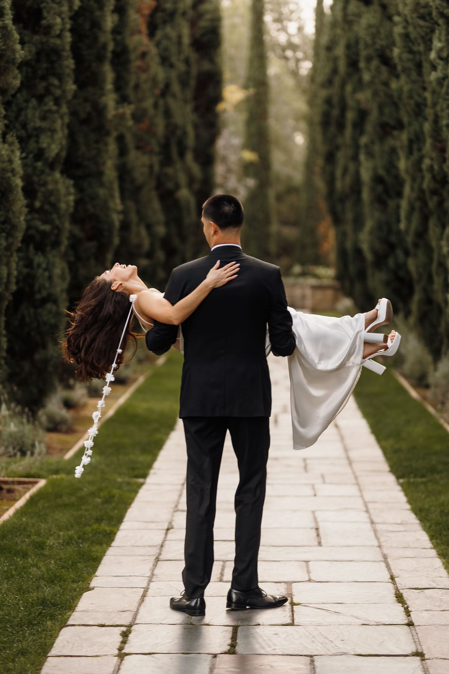 Groom carrying bride in wedding dress along a cypress tree-lined garden pathway.