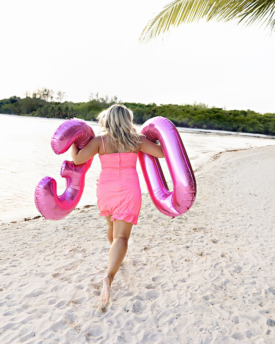 Woman in pink dress running on beach holding large pink number 30 balloons, celebrating her 30th birthday.