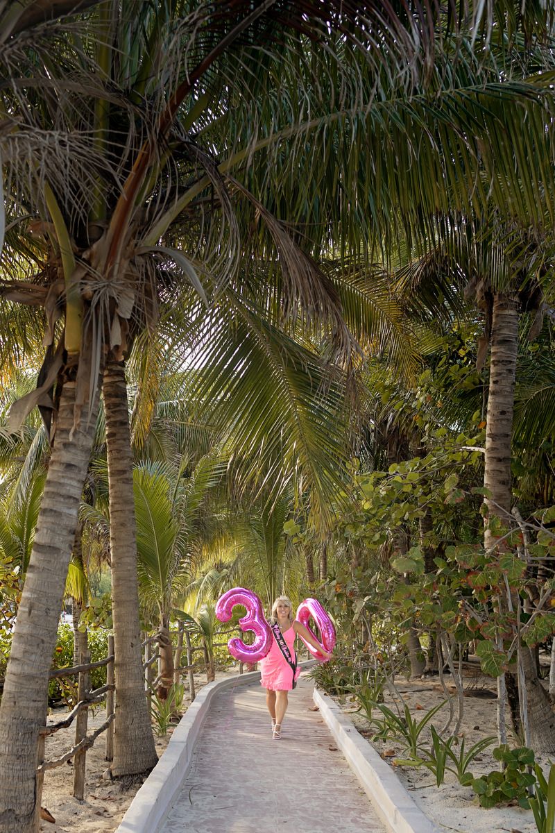 Woman in pink dress walks palm-lined path holding pink number 30 balloons at tropical resort.