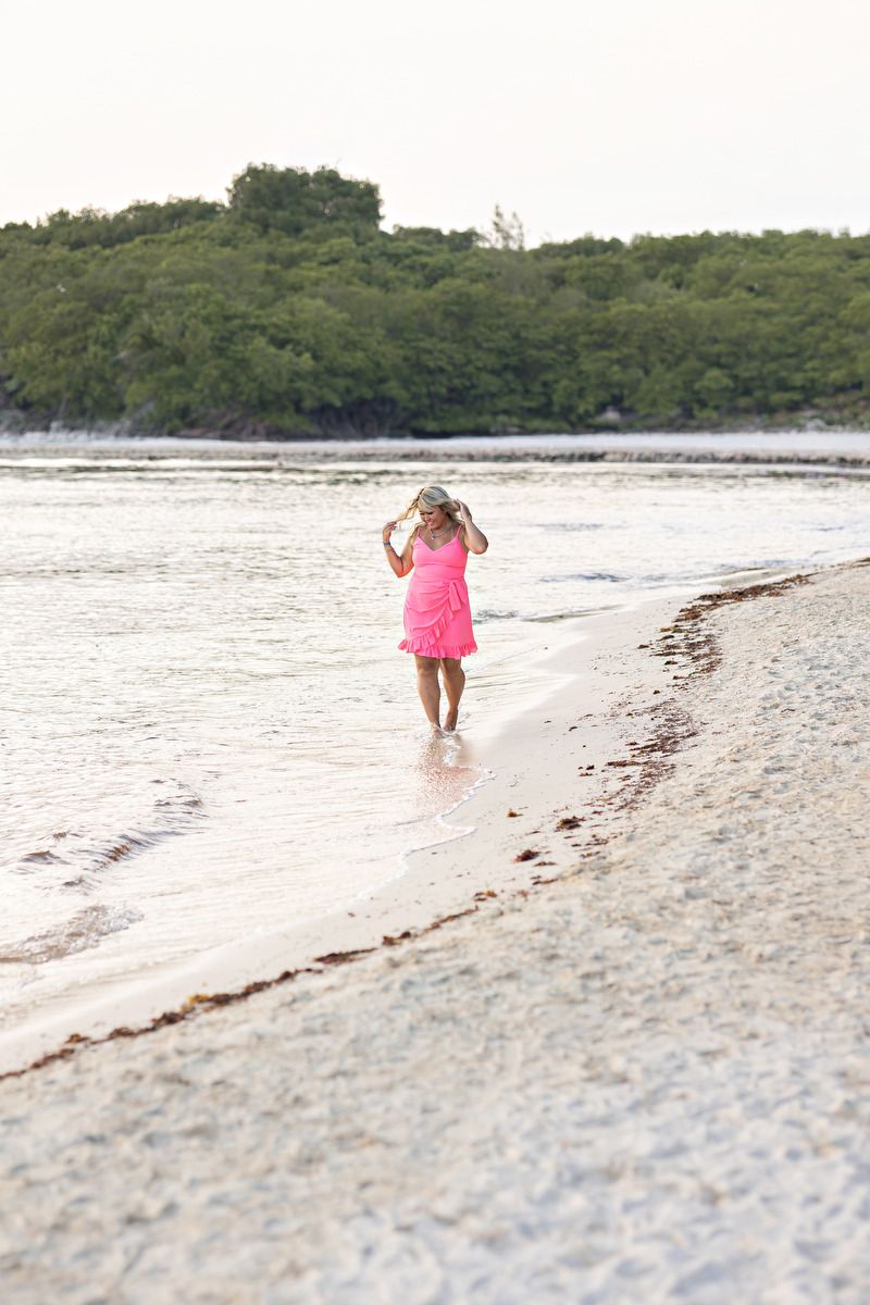 Woman in pink dress walks along serene tropical beach shoreline at sunset.