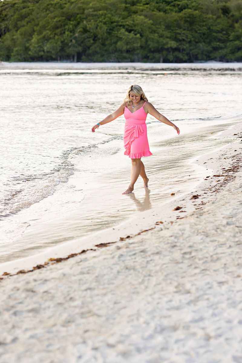 Woman in pink dress walking along a tropical beach shoreline with lush green trees in the background.