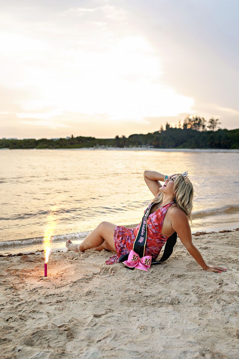 Woman in pink dress with sash sits on sandy beach at sunset near a lit flare.