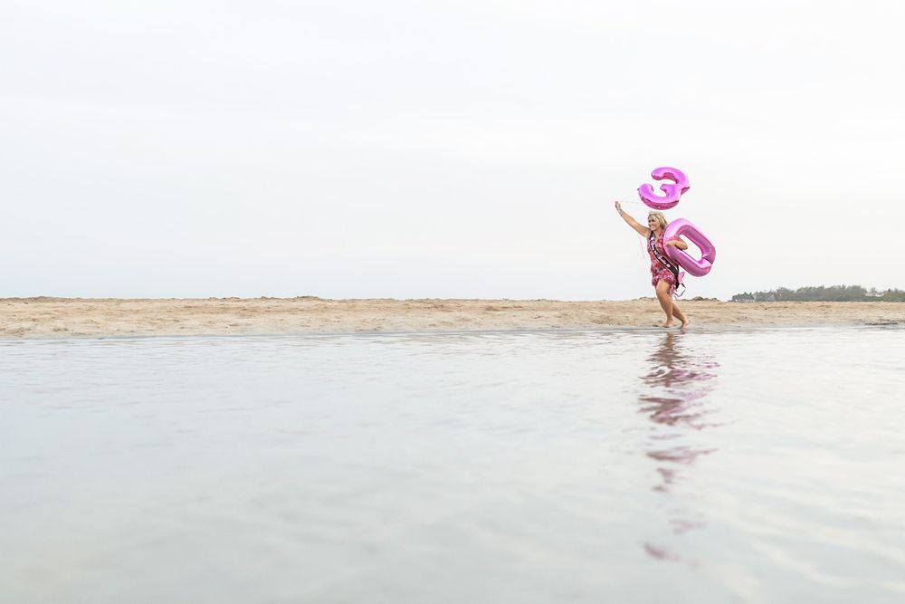 A young girl runs joyfully on a beach carrying pink inflatable rings on a calm, overcast day.