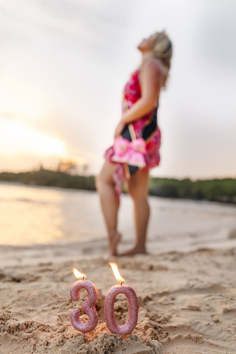 Pink glittery 30th birthday candles lit in beach sand with blurred woman in floral dress at sunset.
