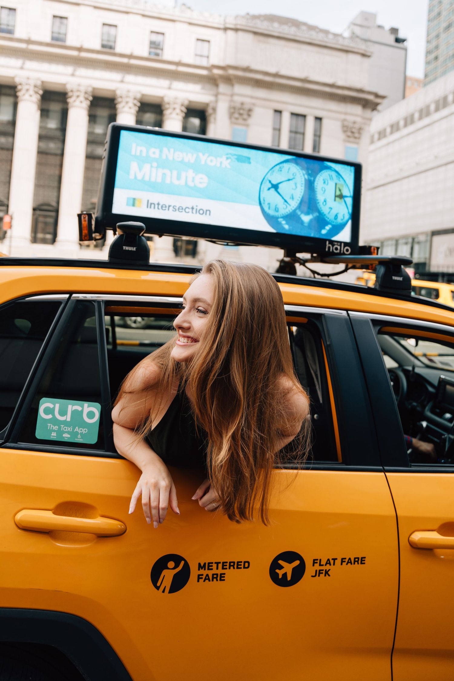 Woman leaning out of yellow NYC taxi window with city buildings and blue sign in background.