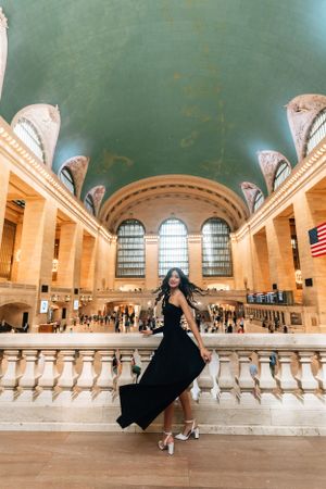 Person in flowing black dress poses dramatically on grand staircase of historic train station.