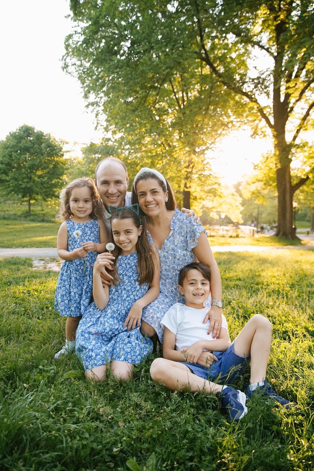 Happy family of five posing together on green grass in a sunlit park, wearing coordinating blue outfits.
