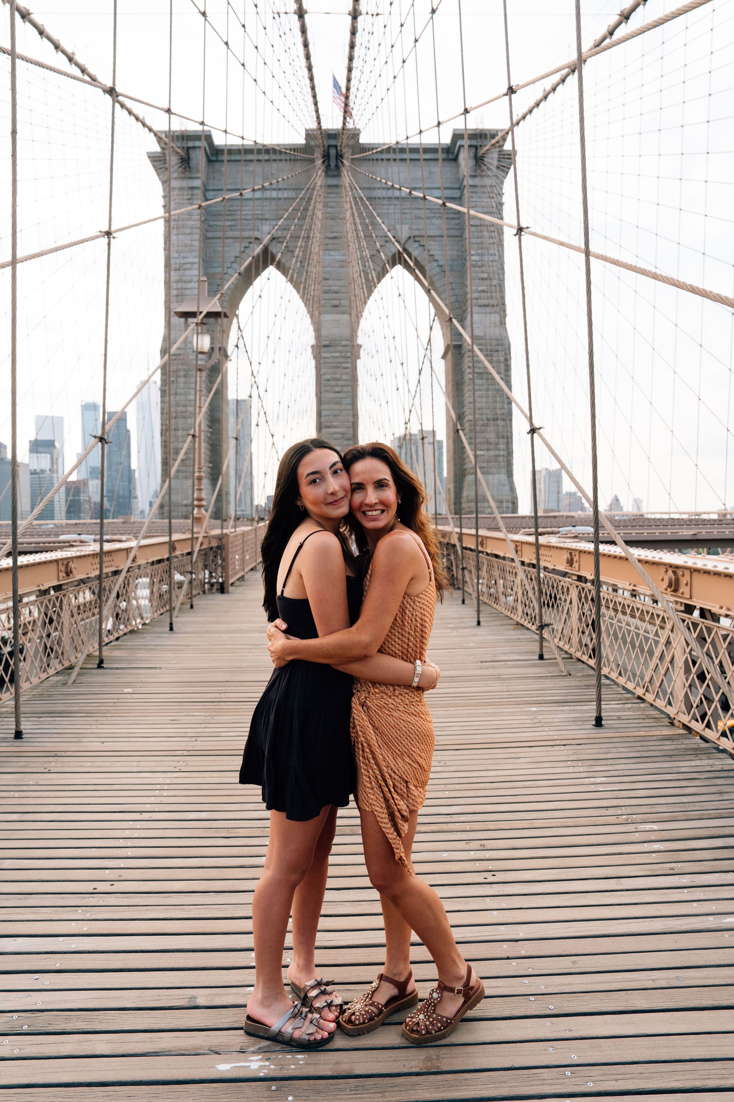Two women hugging and smiling on the Brooklyn Bridge wooden walkway with the iconic tower behind them.