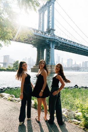 Three women in black outfits pose by the Manhattan Bridge in NYC with the skyline visible behind them.