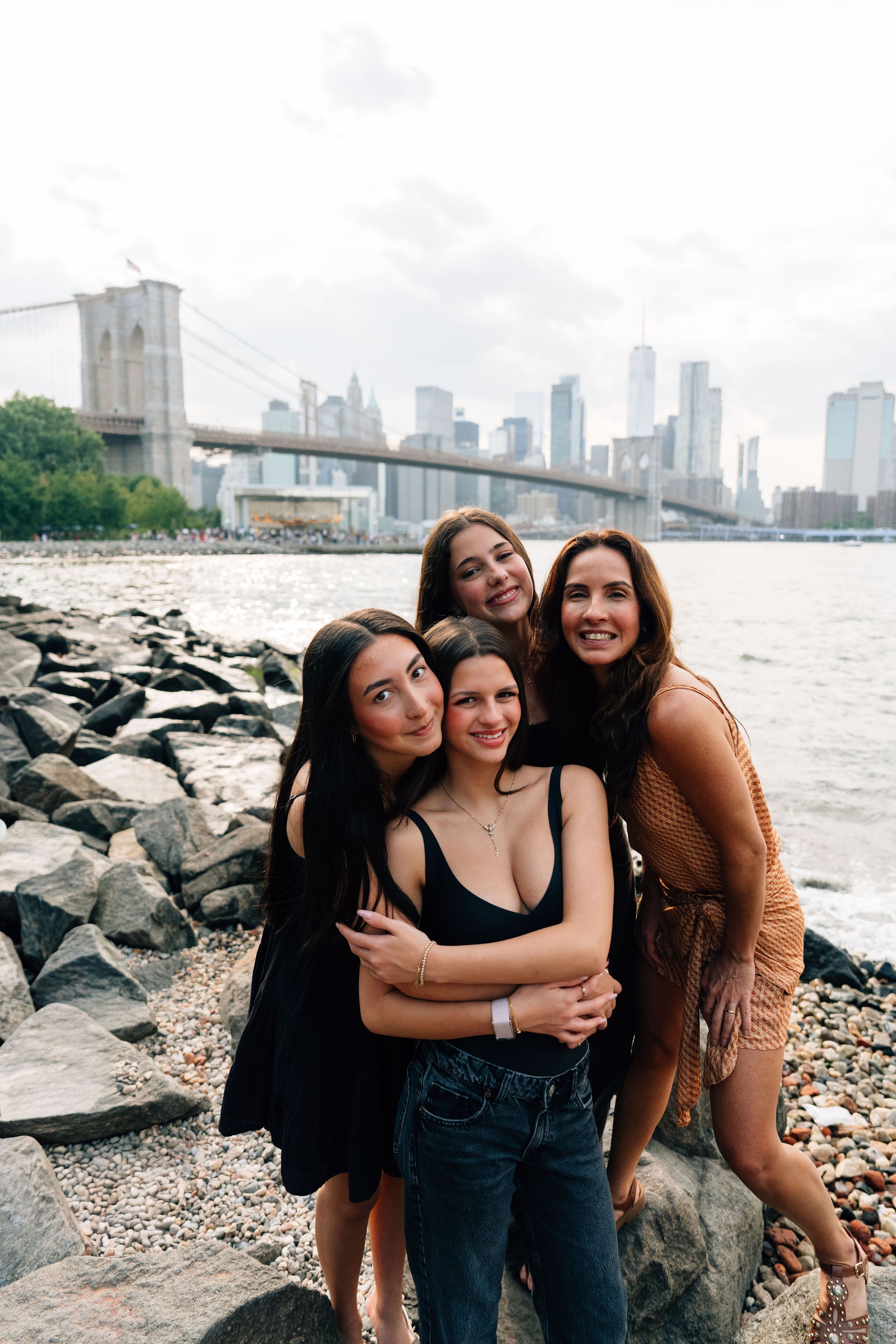 Four women pose together on rocky Brooklyn waterfront with Brooklyn Bridge and NYC skyline in background.