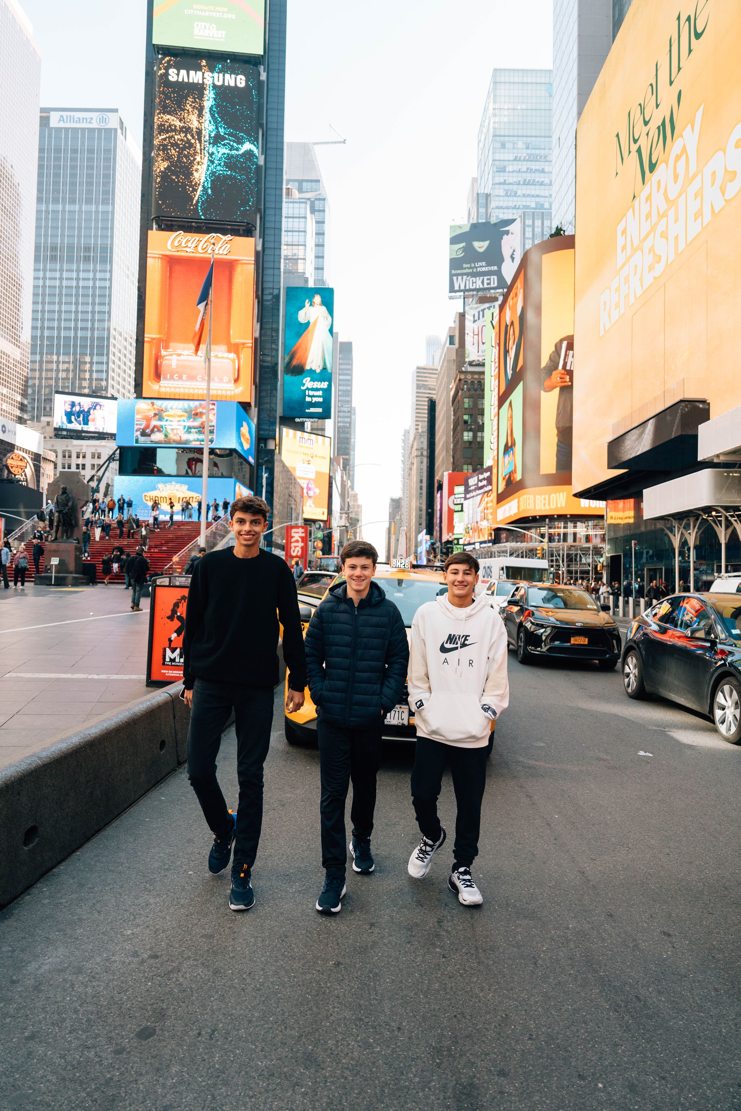 Three young men posing together in Times Square, New York City, surrounded by iconic billboards and busy streets.