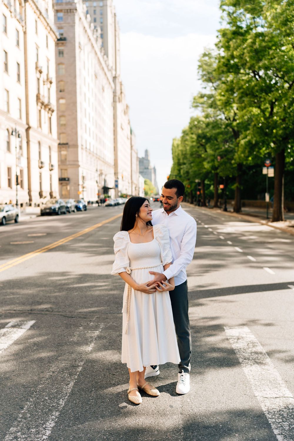 A couple stands together on an empty city street, the woman in a white dress and the man in a white shirt, sharing a tender moment.