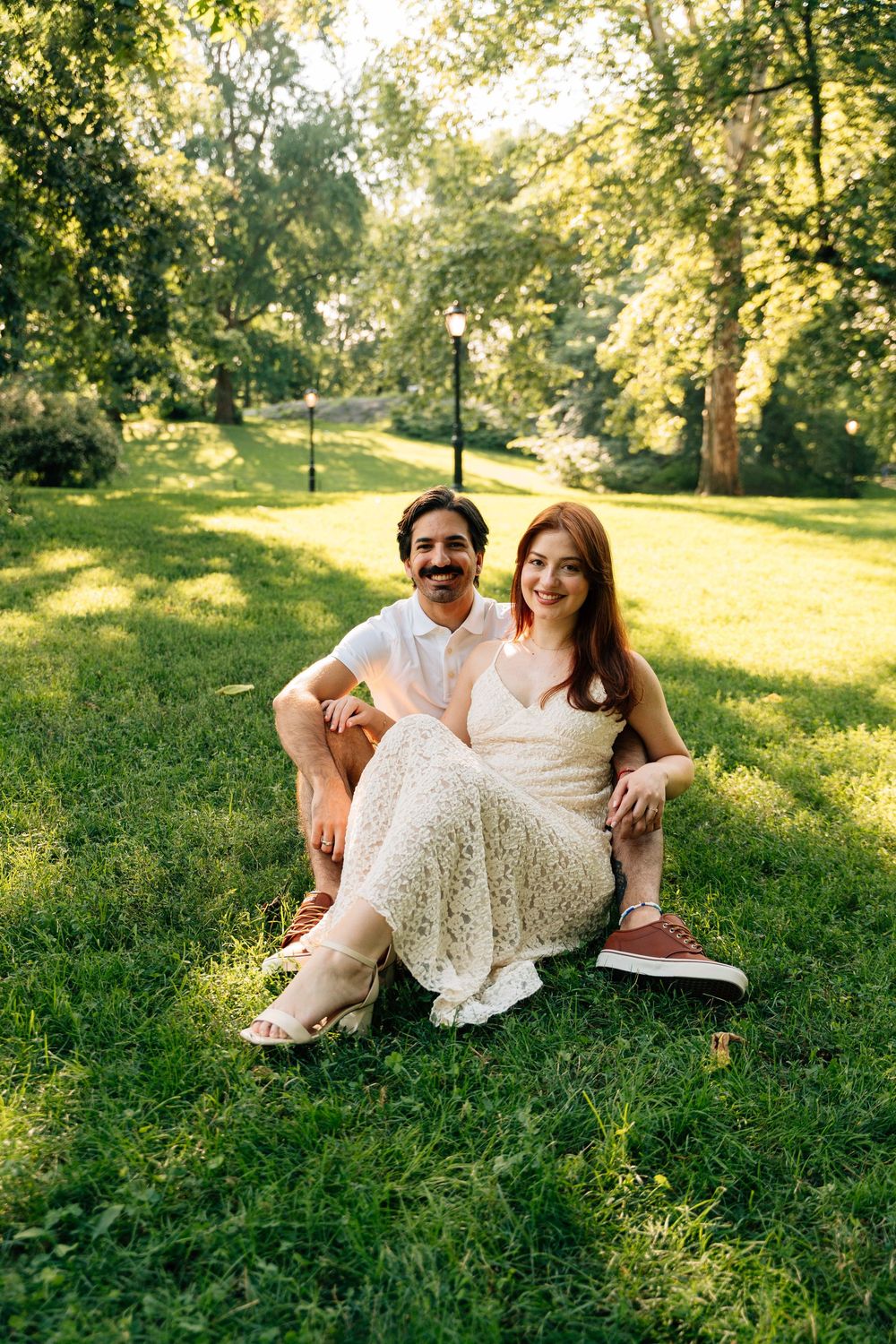 A couple sits together on green grass in a sunny park, smiling at the camera in casual summer attire.