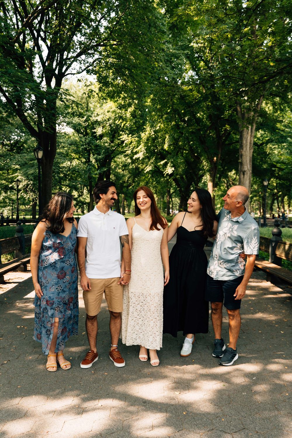Five people smiling and walking together on a shaded park path surrounded by lush green trees on a sunny day.