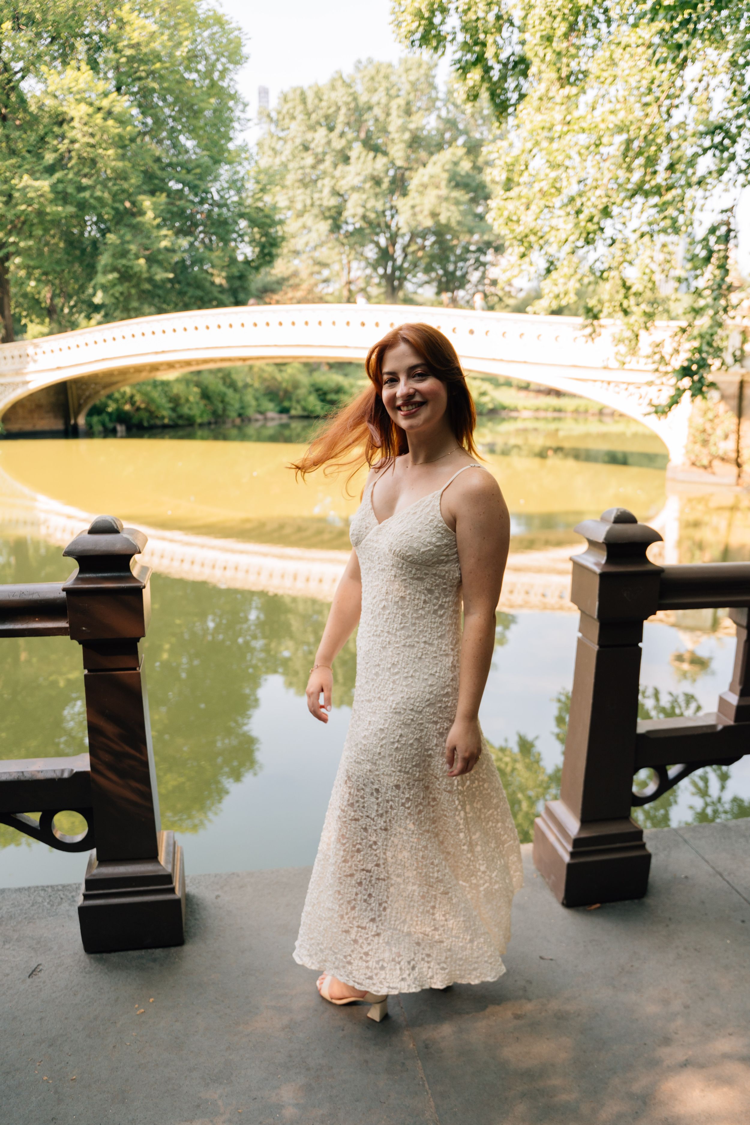 A smiling woman in a white lace dress poses near a pond with Bow Bridge in Central Park behind her.