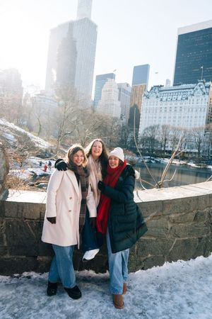 Three women smiling in snowy Central Park, NYC, wearing winter coats with skyline behind them.