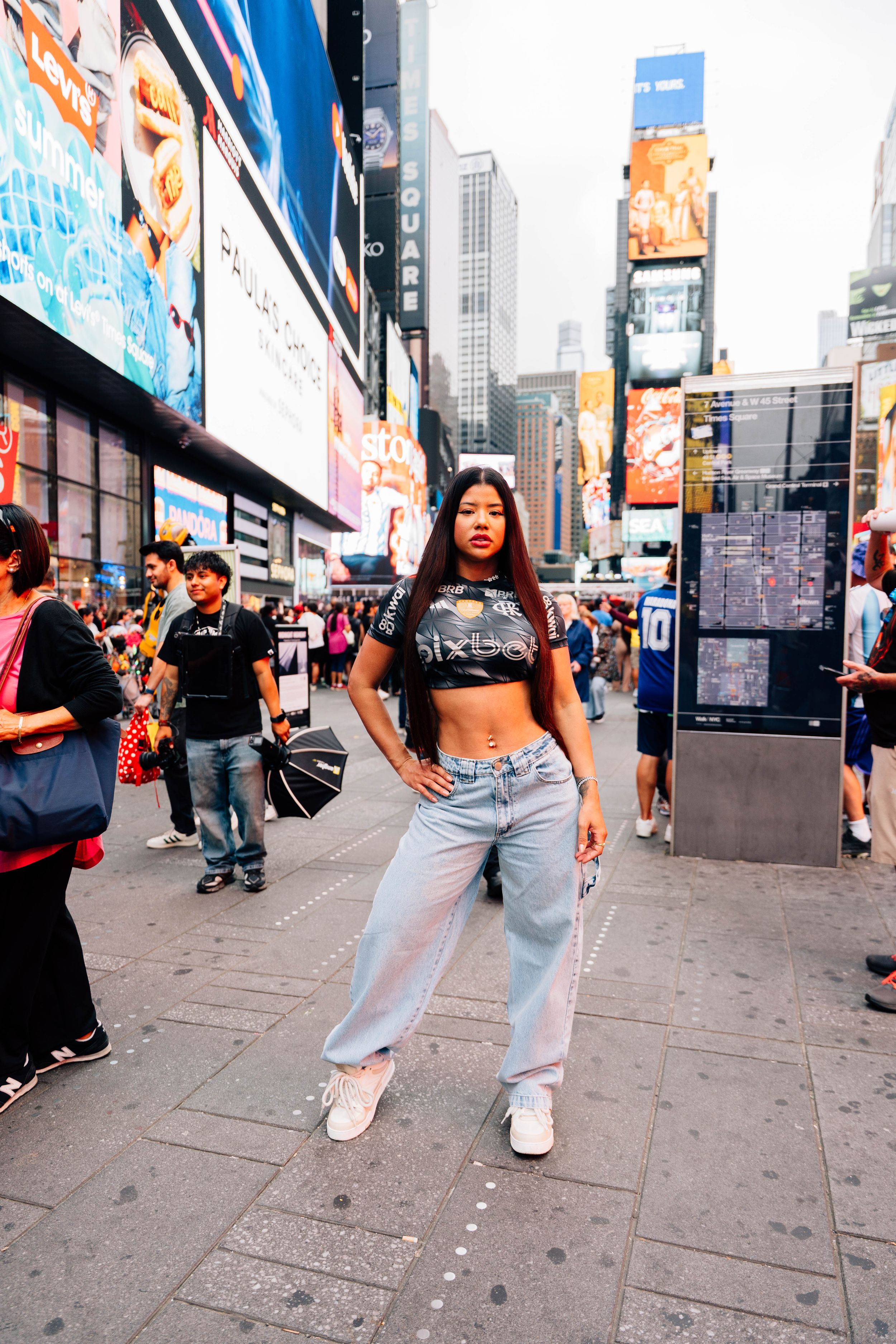 Young woman in crop top and baggy jeans posing confidently in Times Square, NYC, surrounded by crowds and billboards.