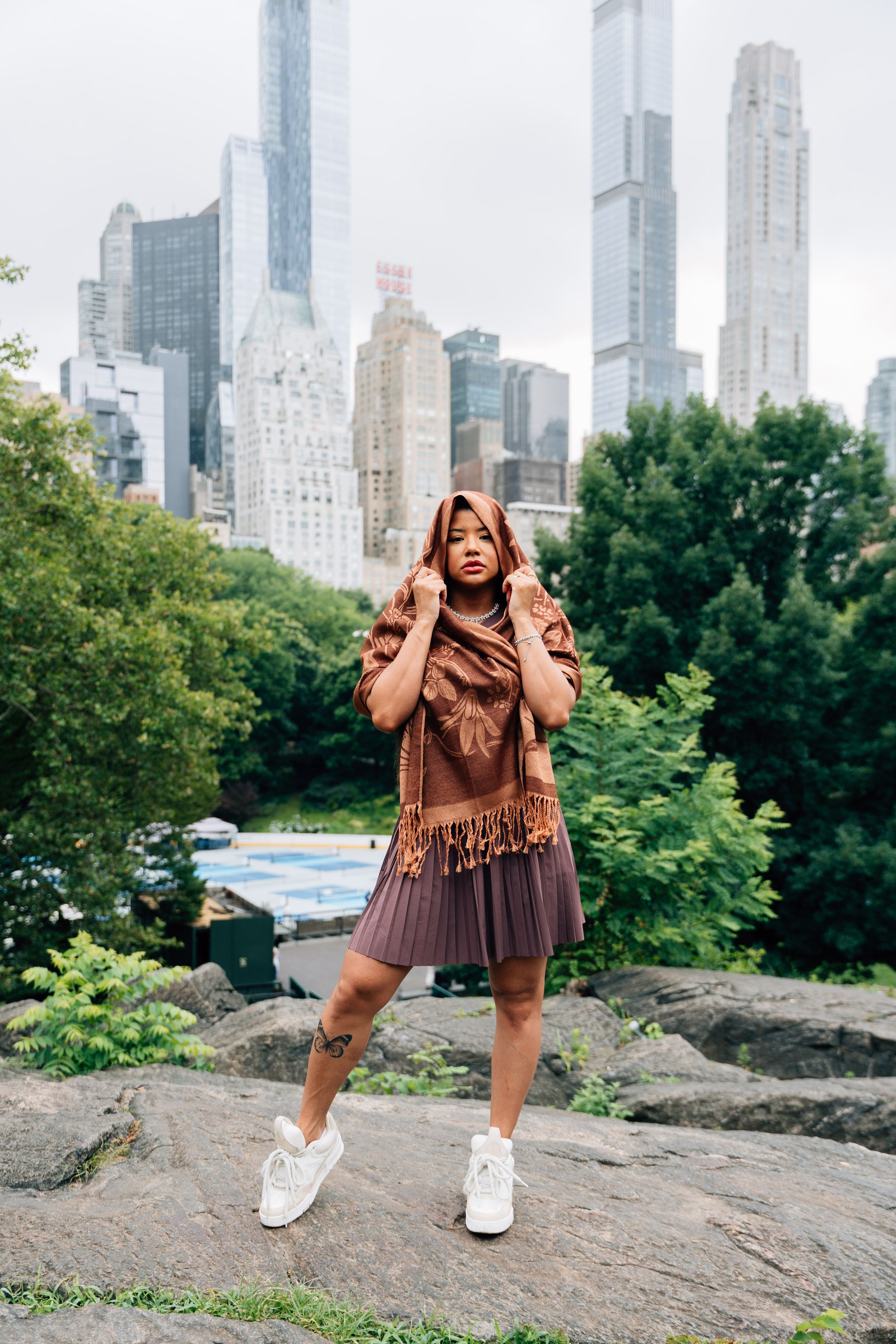 Young woman in brown dress and scarf posing on rocks in Central Park with NYC skyline in background.
