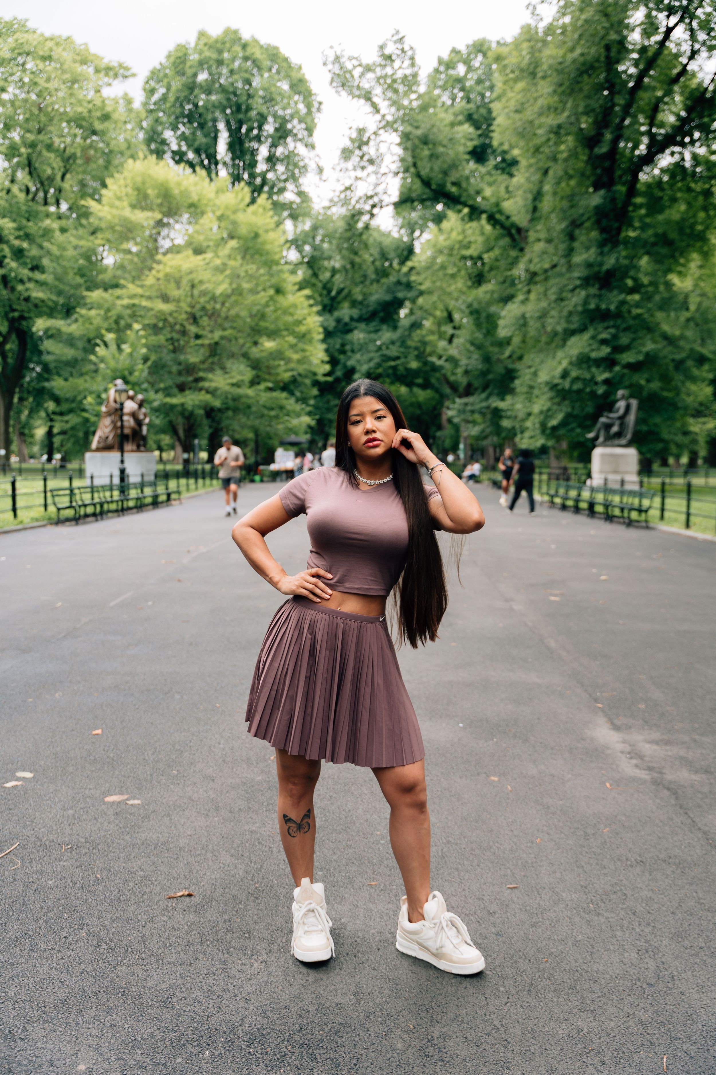 Young woman in matching brown crop top and pleated skirt poses confidently in a lush green park pathway.